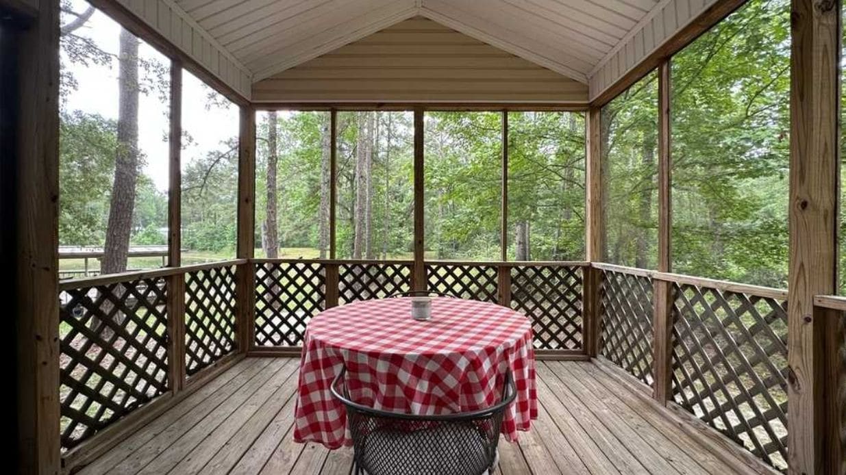 A covered outdoor area with a table and chairs, surrounded by a wooden deck and lattice railing, overlooking a forested area.