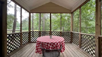 A covered outdoor area with a table and chairs, surrounded by a wooden deck and lattice railing, overlooking a forested area.