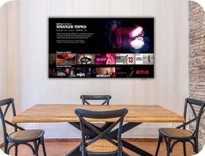 Eine wandmontierte Anzeige zeigt eine Netflix-Originalserie, umgeben von einem Essbereich mit einem Holztisch und schwarzen Stühlen.