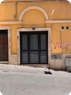 Ein Gebäude mit gelber Fassade, einer schwarzen Tür mit Fensterfeldern und der Hausnummer 27 an der Wand.