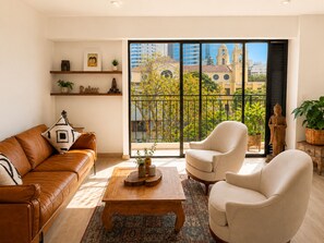 A living room with a brown leather sofa, a beige armchair, a wooden coffee table, and a large window with a view of buildings and trees. - Deluxe Retreat Steps from Larcomar (Miraflores)