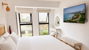 A bedroom with a large bed, a bedside table, a bench, a mirror, and a window with a view of a coastal landscape. - Deluxe Retreat Steps from Larcomar (Miraflores)