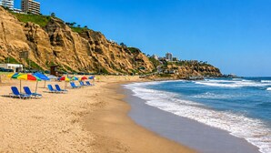 A beach with blue lounge chairs and umbrellas, a rocky cliff, and buildings in the background. - Deluxe Retreat Steps from Larcomar (Miraflores)