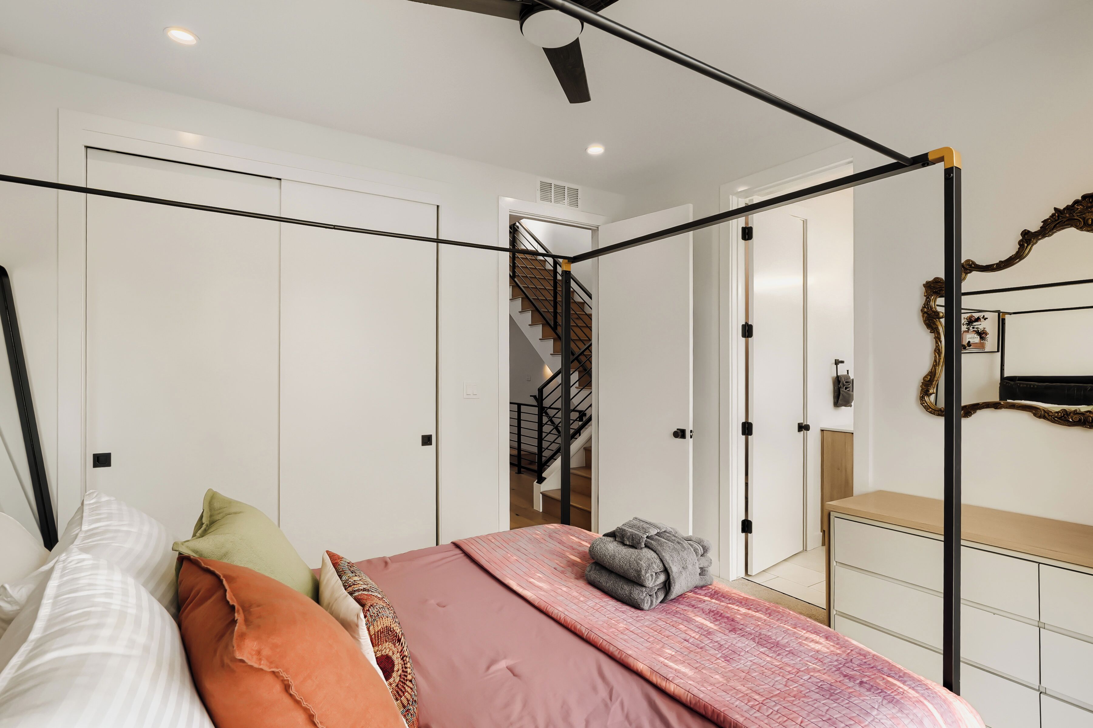 A bedroom with a four-poster bed, a chest of drawers, a mirror, and a staircase leading to another room.