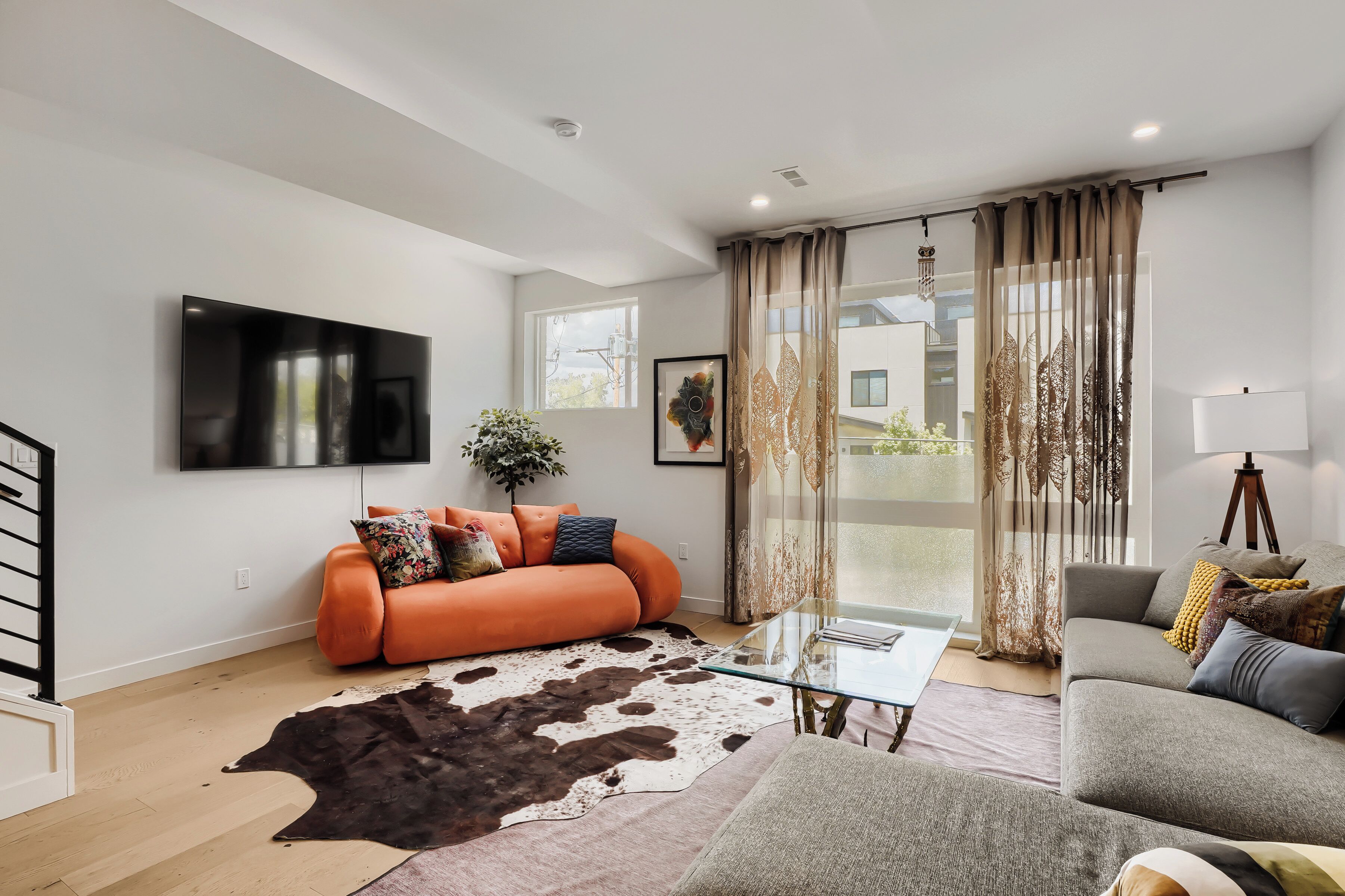 A modern living room with a sofa, a glass coffee table, a mounted TV, and a large window with curtains.