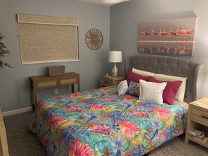 Ein Schlafzimmer mit blumigem Bettüberwurf, einem grau gepolsterten Kopfteil, einem Nachttisch mit Lampe und einem Wandbild von Flamingos.