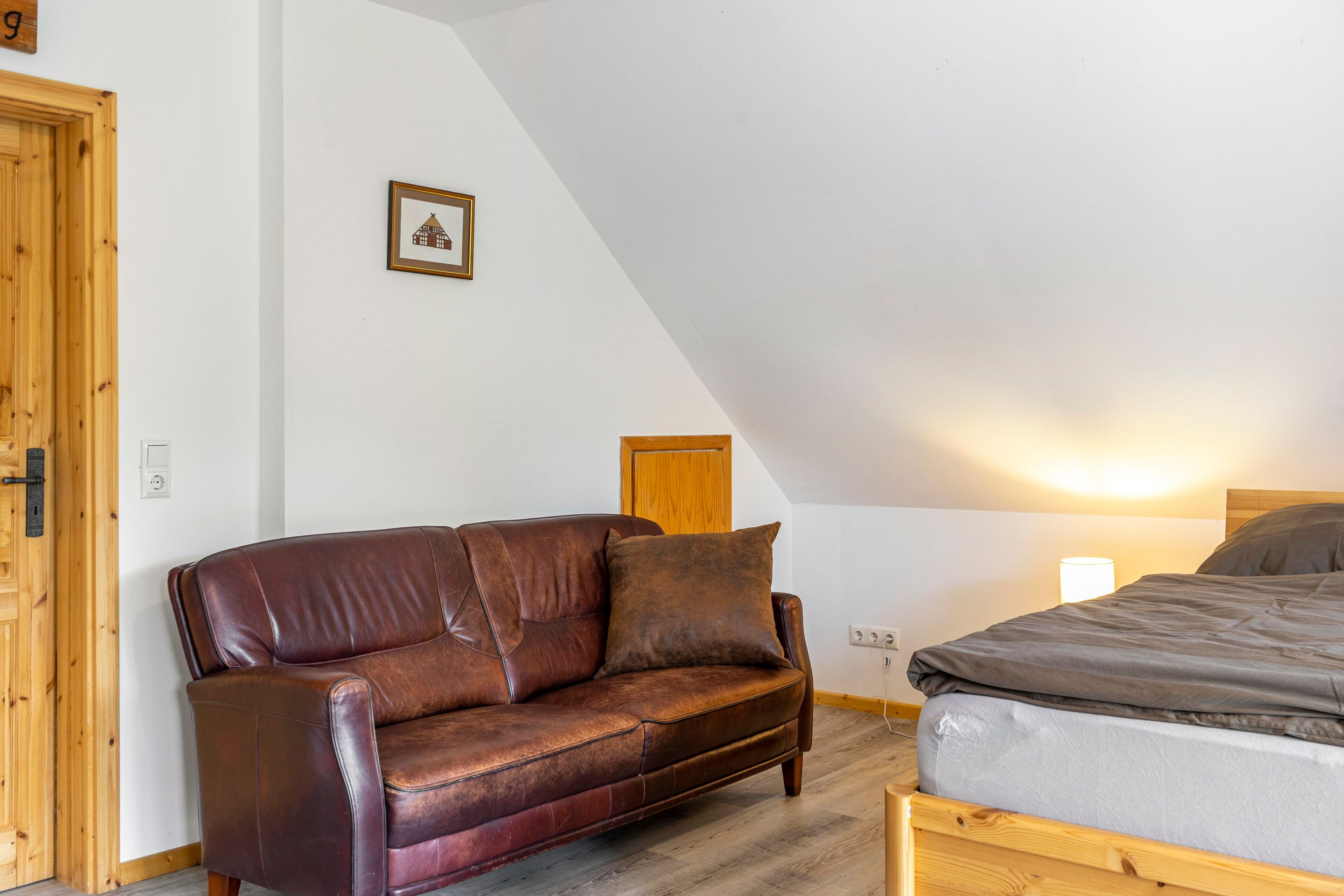Ein Zimmer mit einem braunen Ledersofa, einem hölzernen Bett, einem gerahmten Bild an der Wand und einer Holztür.