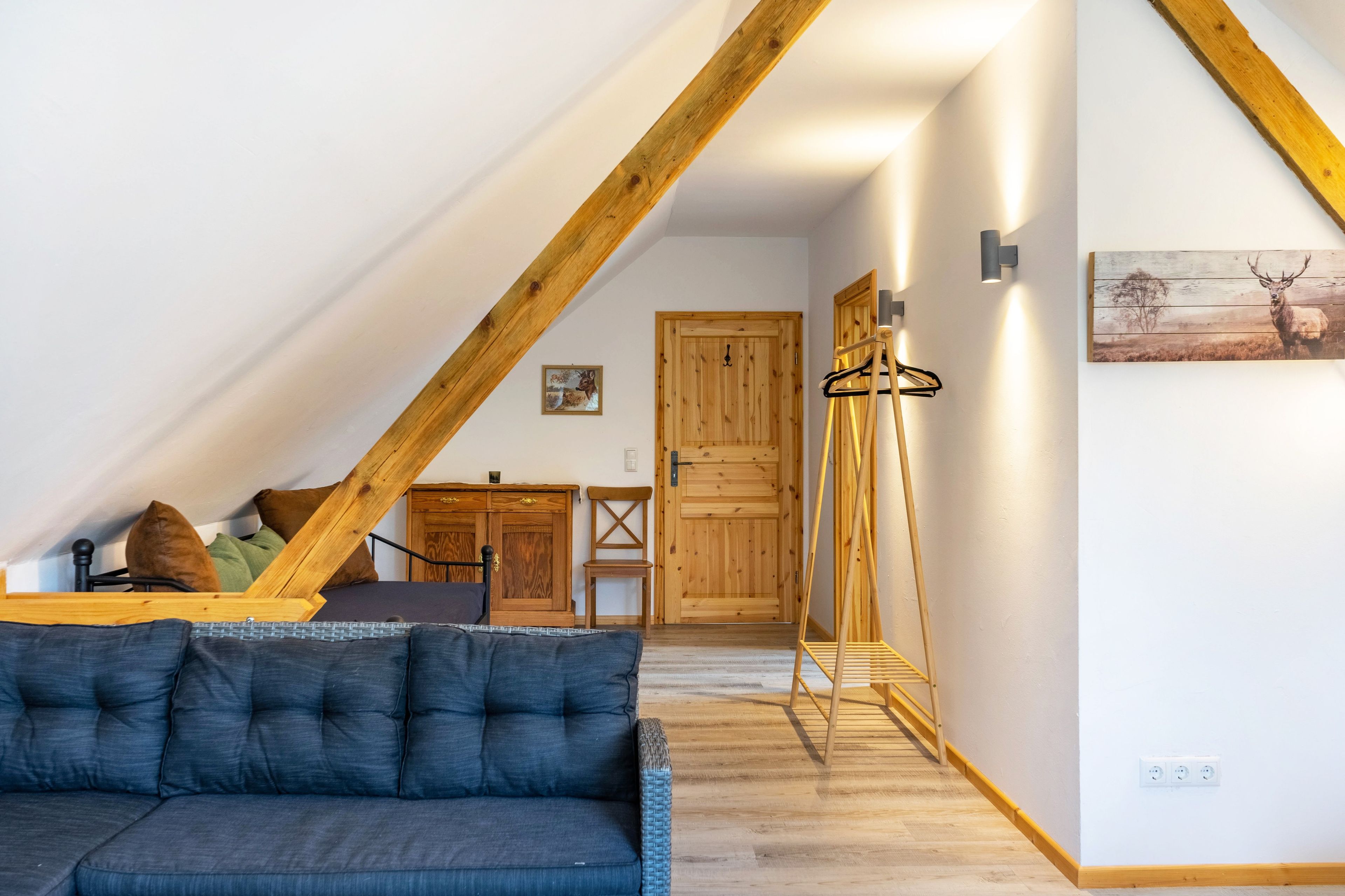 Una habitación con techo inclinado, un sofá, un armario de madera, una puerta y una lámpara fijada a la pared.