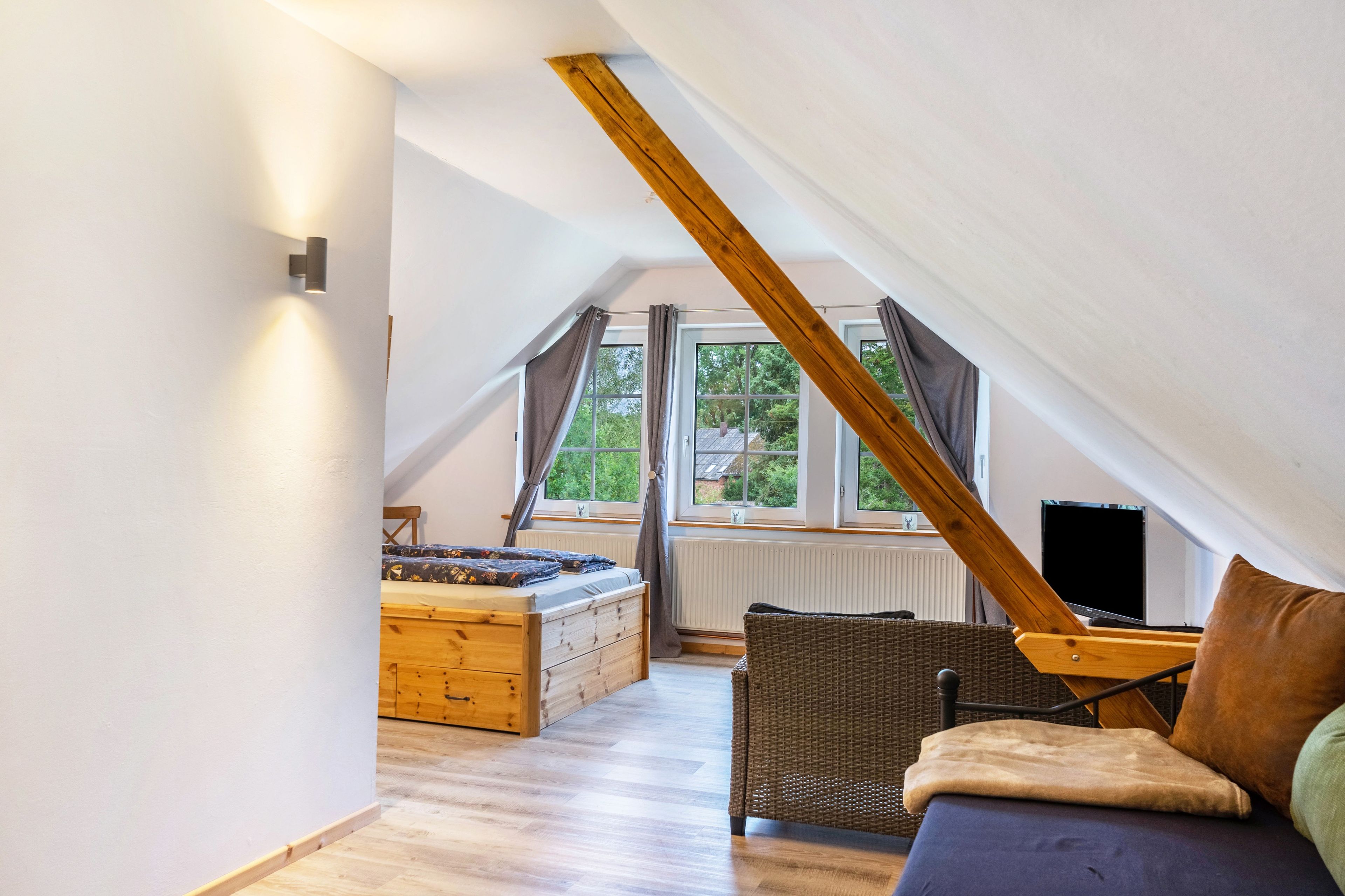 A bedroom with a sloped ceiling, wooden beams, a bed, a wicker chair, and a window with curtains.