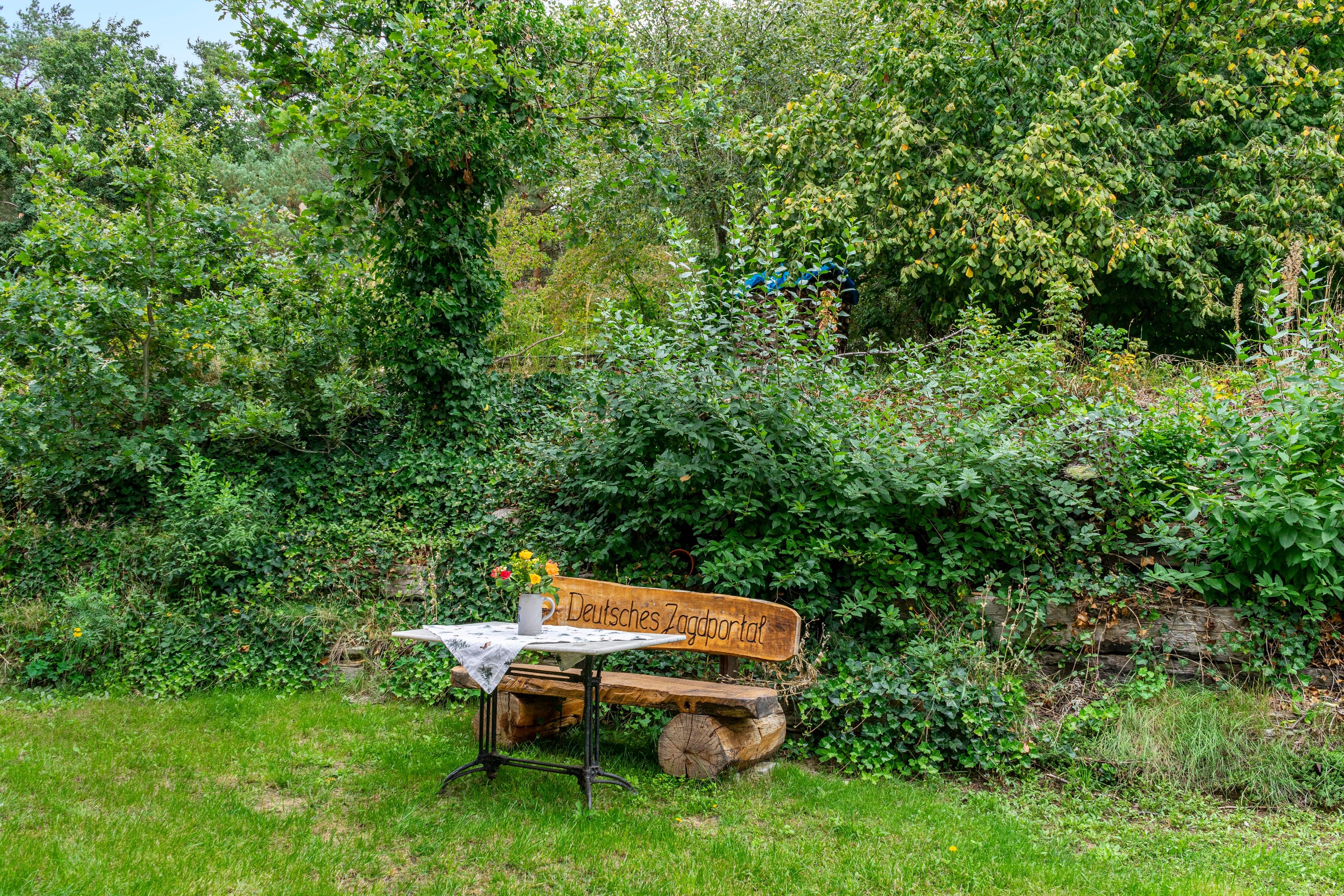 Een rustieke houten bank met een bord waarop 'Deutsches Zagportal' staat, in een weelderige tuin.