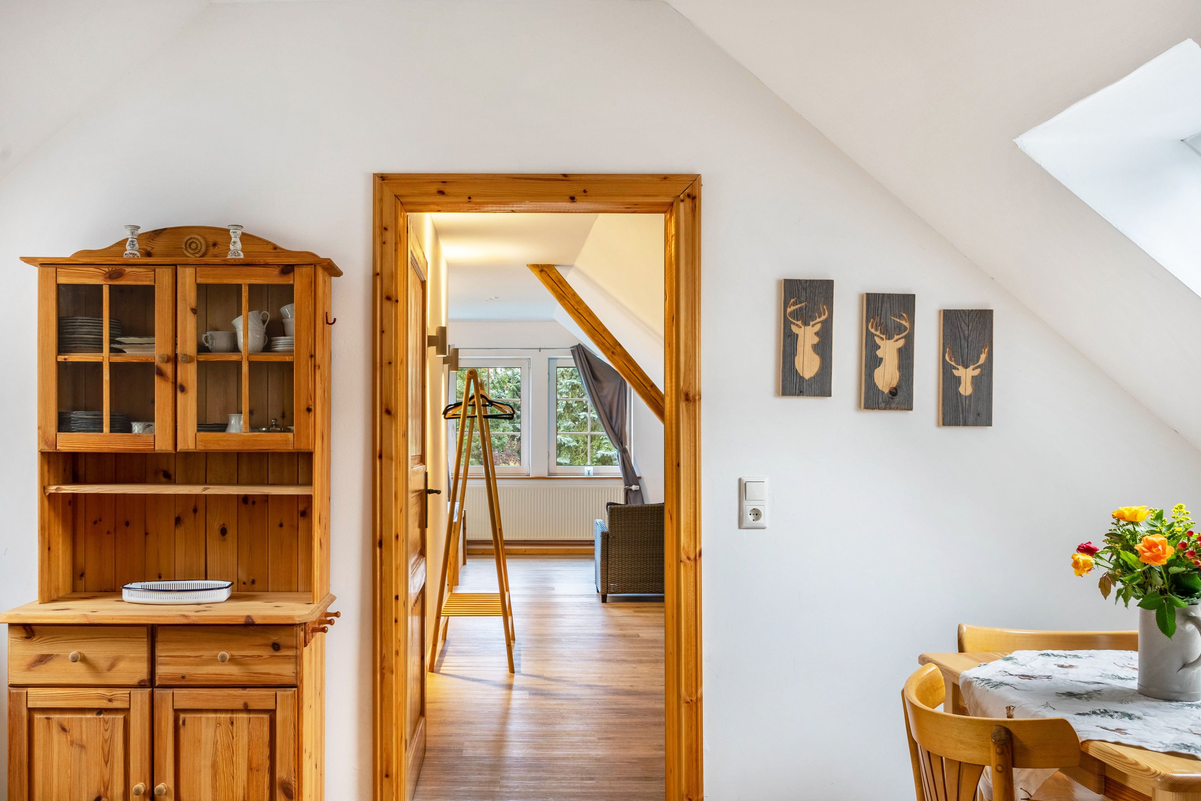 Une pièce avec un meuble en bois, une table ornée d’un vase de fleurs et une vue sur une maison, visible à travers une porte.
