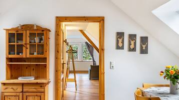 Une pièce avec un meuble en bois, une table ornée d’un vase de fleurs et une vue sur une maison, visible à travers une porte.
