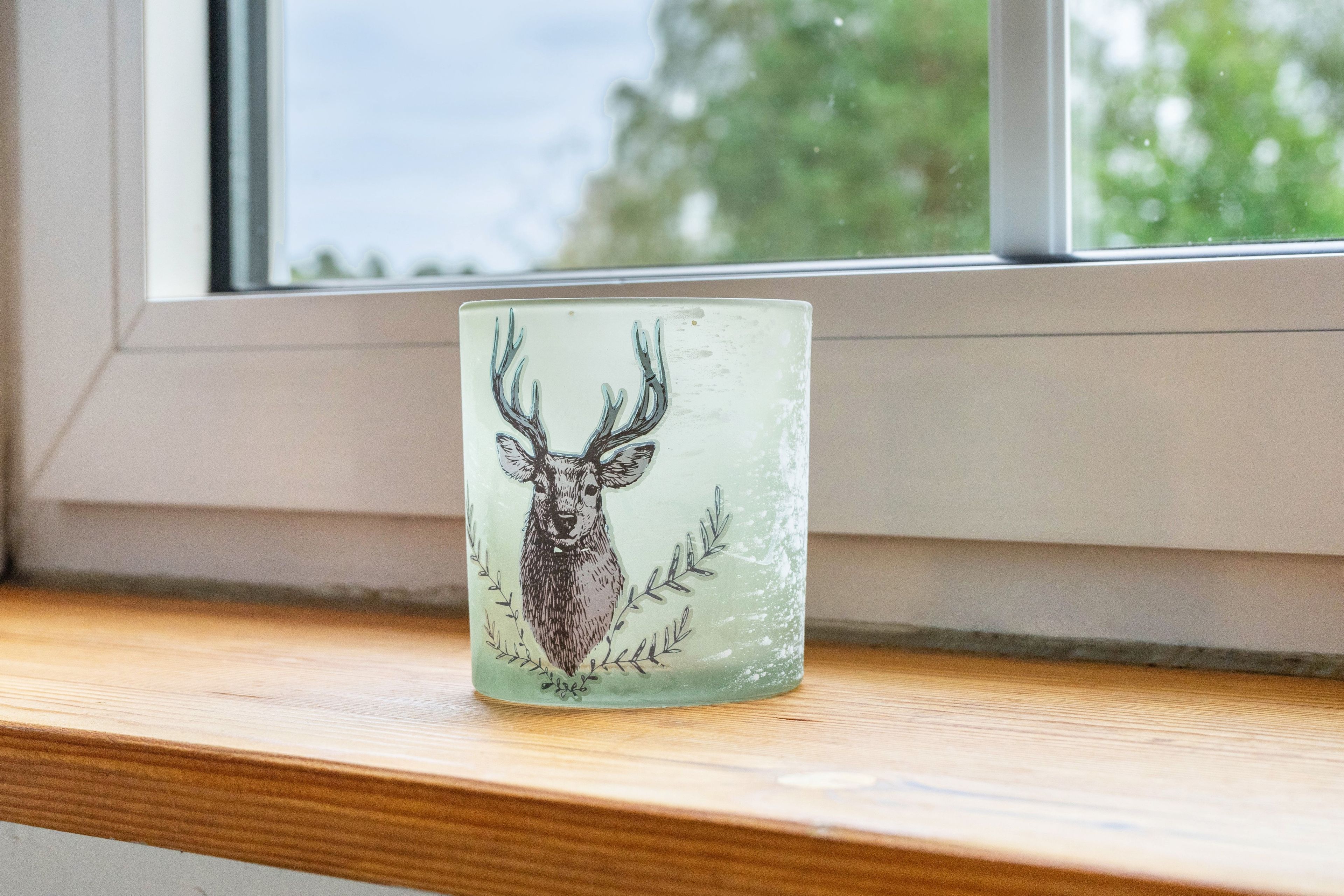 A glass mug with a deer illustration on a wooden ledge by a window.
