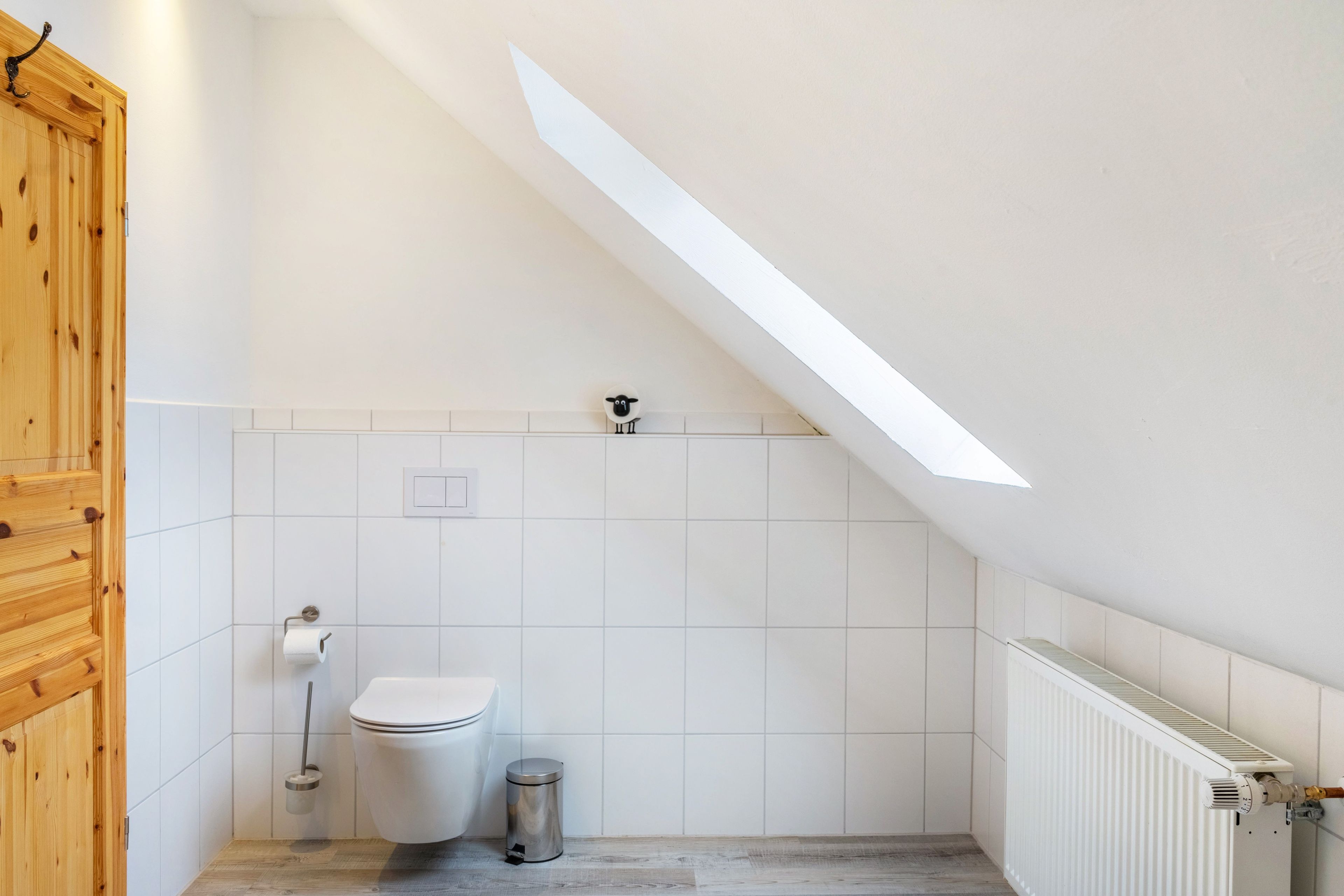 Une petite salle de bain aux murs blancs, avec un plafond en pente, des toilettes, une poubelle et un radiateur.