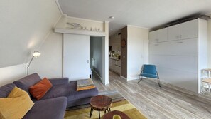 Ein modernes Wohnzimmer mit einer Couch, einem Couchtisch und einem blauen Sessel. Ein Küchenbereich mit Schränken und einer Arbeitsplatte ist durch eine offene Tür sichtbar.