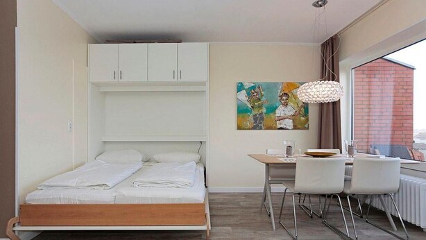 A bedroom with a bed, a desk, and a chair. There is a painting on the wall and a window with a view. - Apartment Vogelsang in Borkum (Borkum)