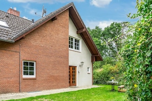 A two-story house with a brick facade, a tiled roof, and a small garden area with a table and chairs.