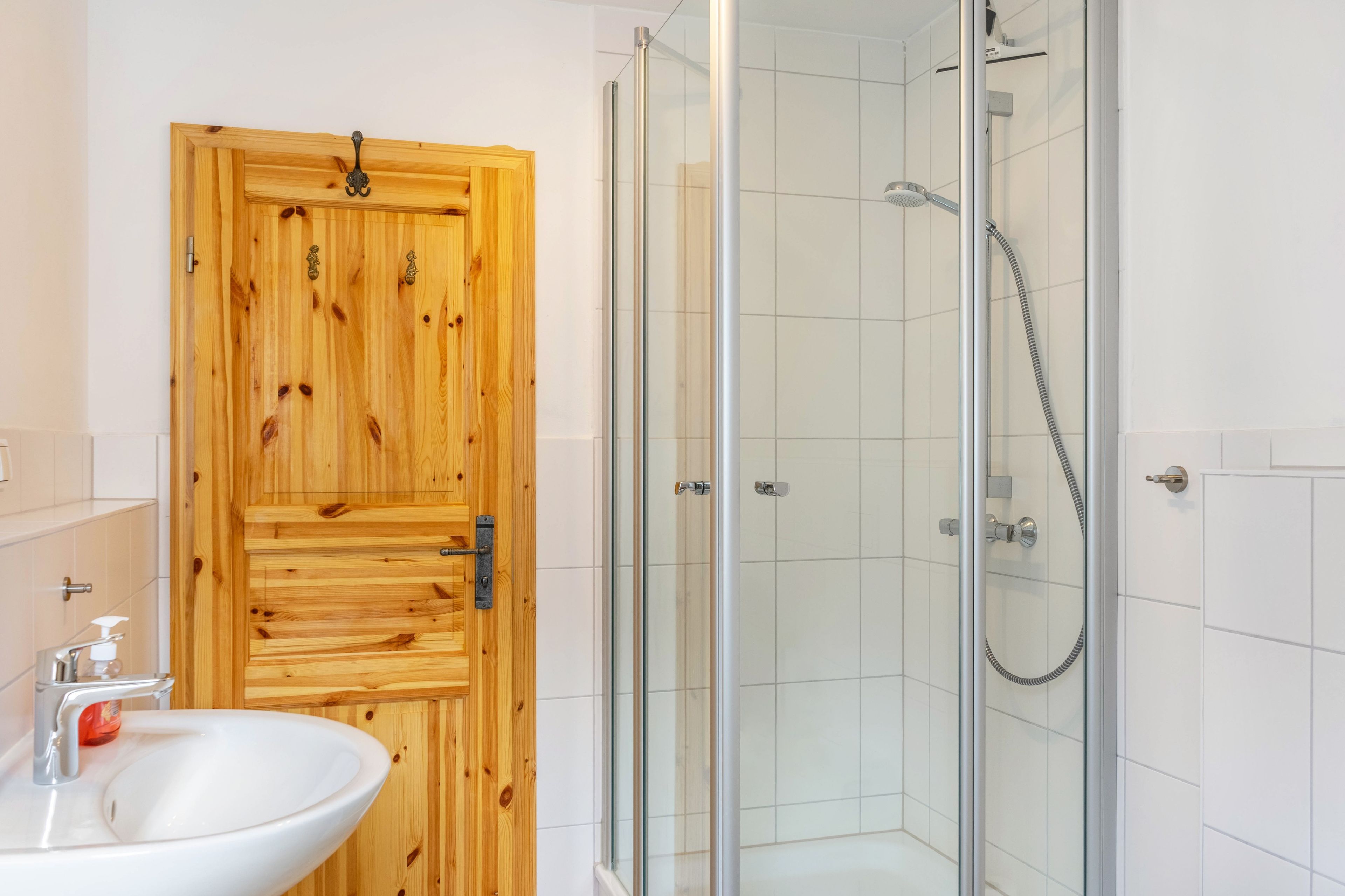 Ein Badezimmer mit Holztür, Duschkabine aus Glas und Waschbecken in Weiß.