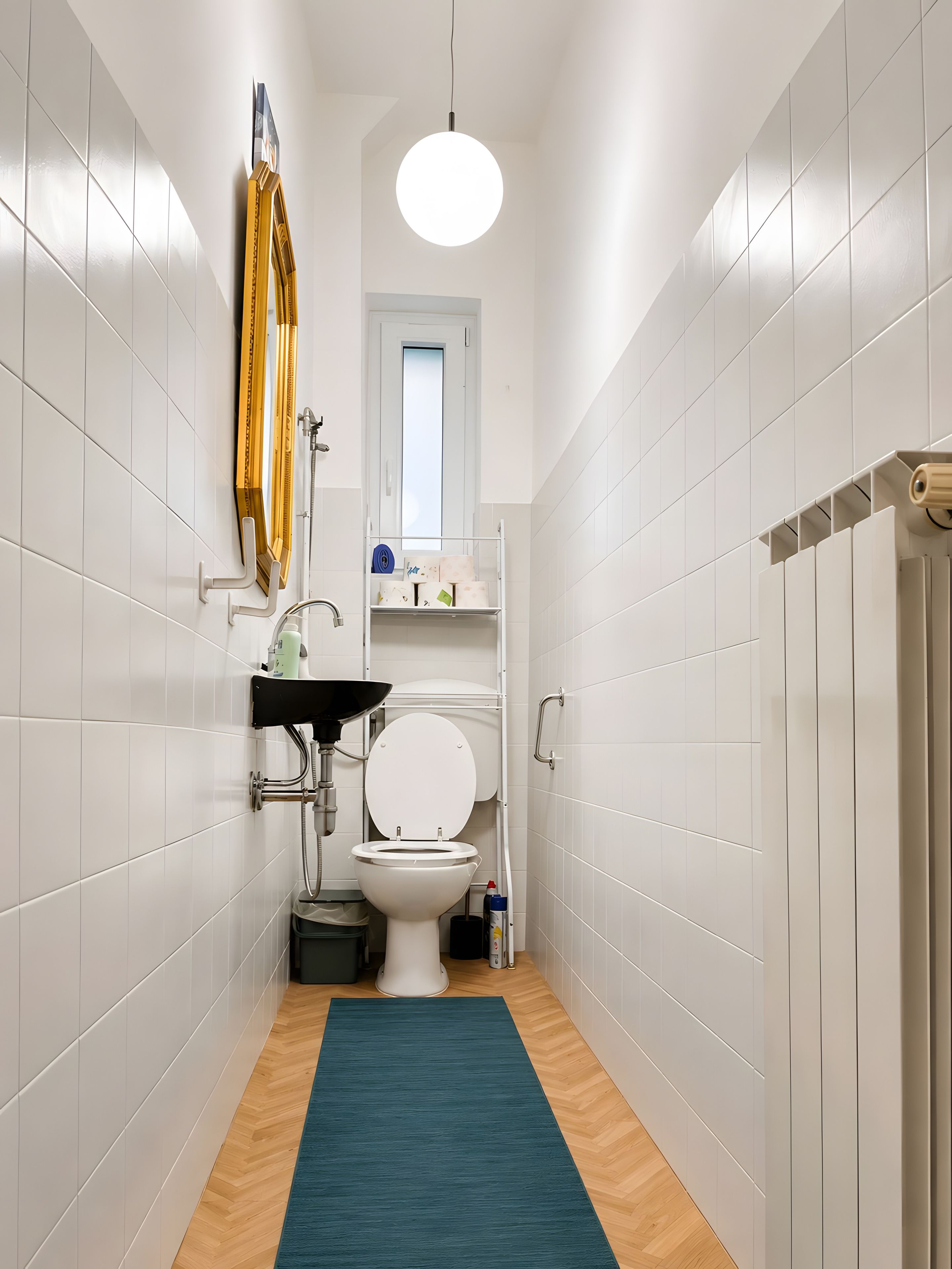 A narrow bathroom with white tiled walls, a wooden floor, a blue rug, a toilet, a sink, and a shower.