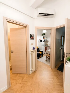 Un couloir doté d’un plancher en bois, d’un climatiseur blanc et d’une porte donnant sur une pièce où se trouvent un lit et une plante.