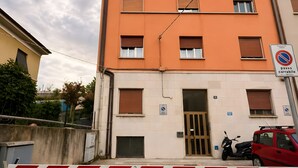 Un immeuble à plusieurs étages doté d’un parking sécurisé par une barrière, avec une voiture rouge et un scooter garés à l’extérieur.