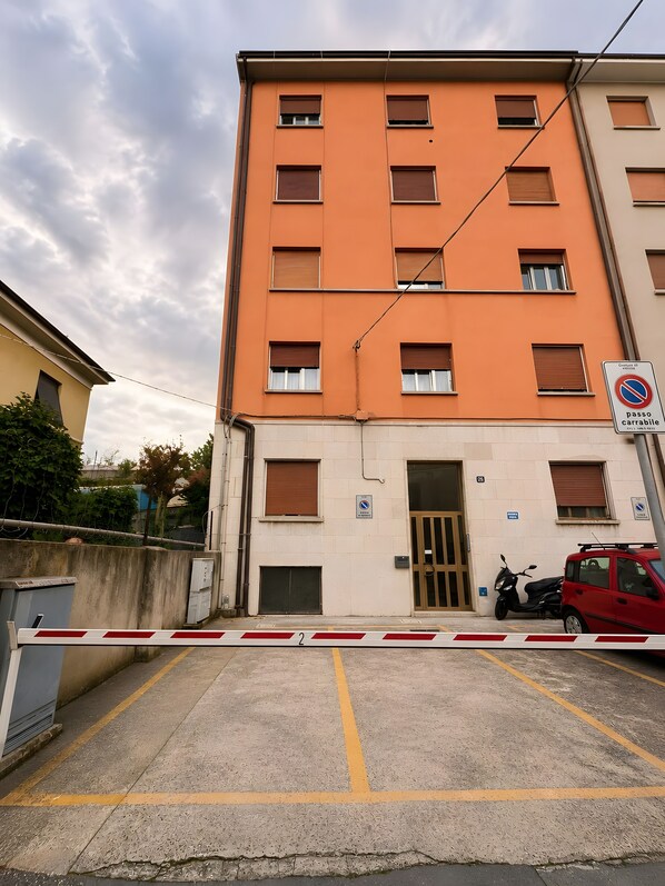 Un immeuble à plusieurs étages doté d’un parking sécurisé par une barrière, avec une voiture rouge et un scooter garés à l’extérieur.