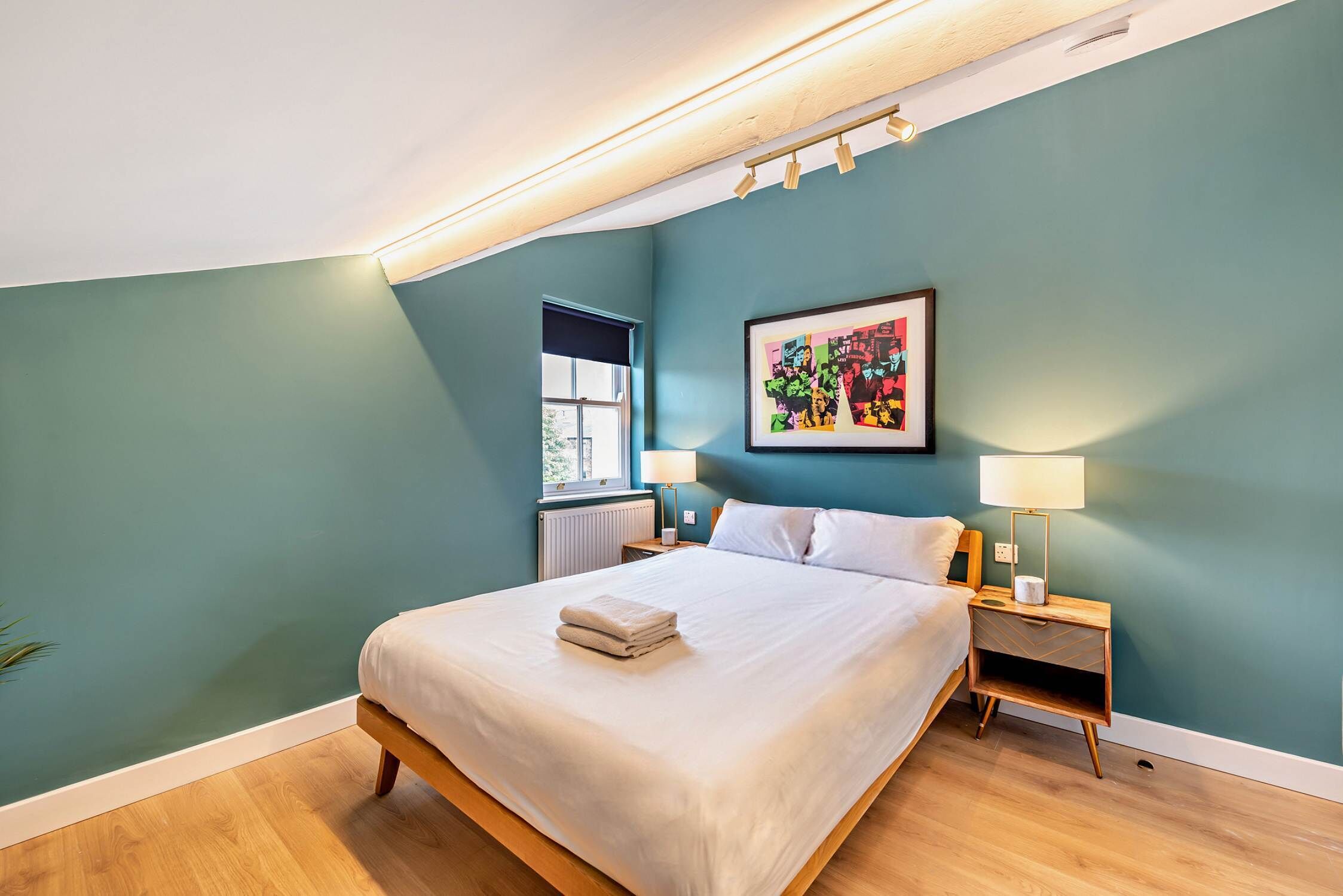 A bedroom with a bed, two bedside tables, a window with a view, and a colorful artwork on the wall.