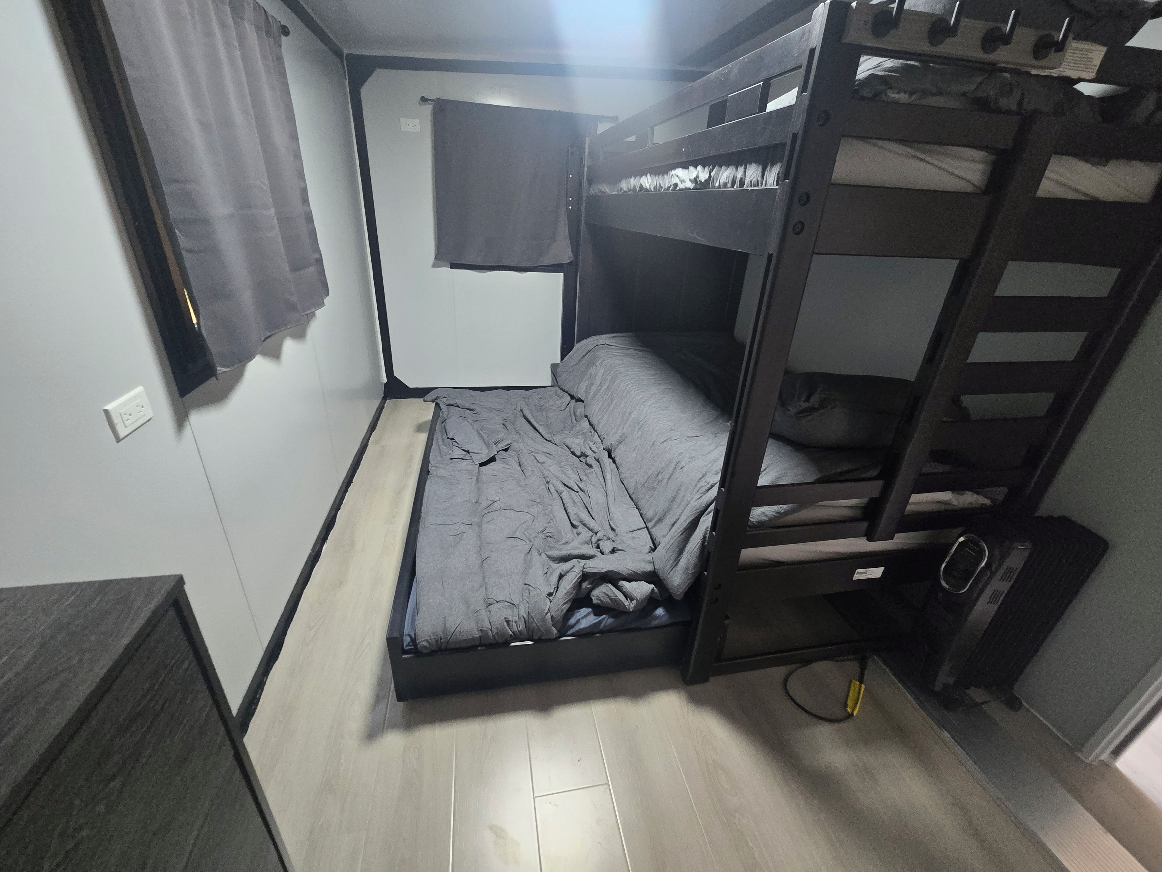 Une chambre avec un lit superposé, une fenêtre, un rideau et une prise de courant au mur.