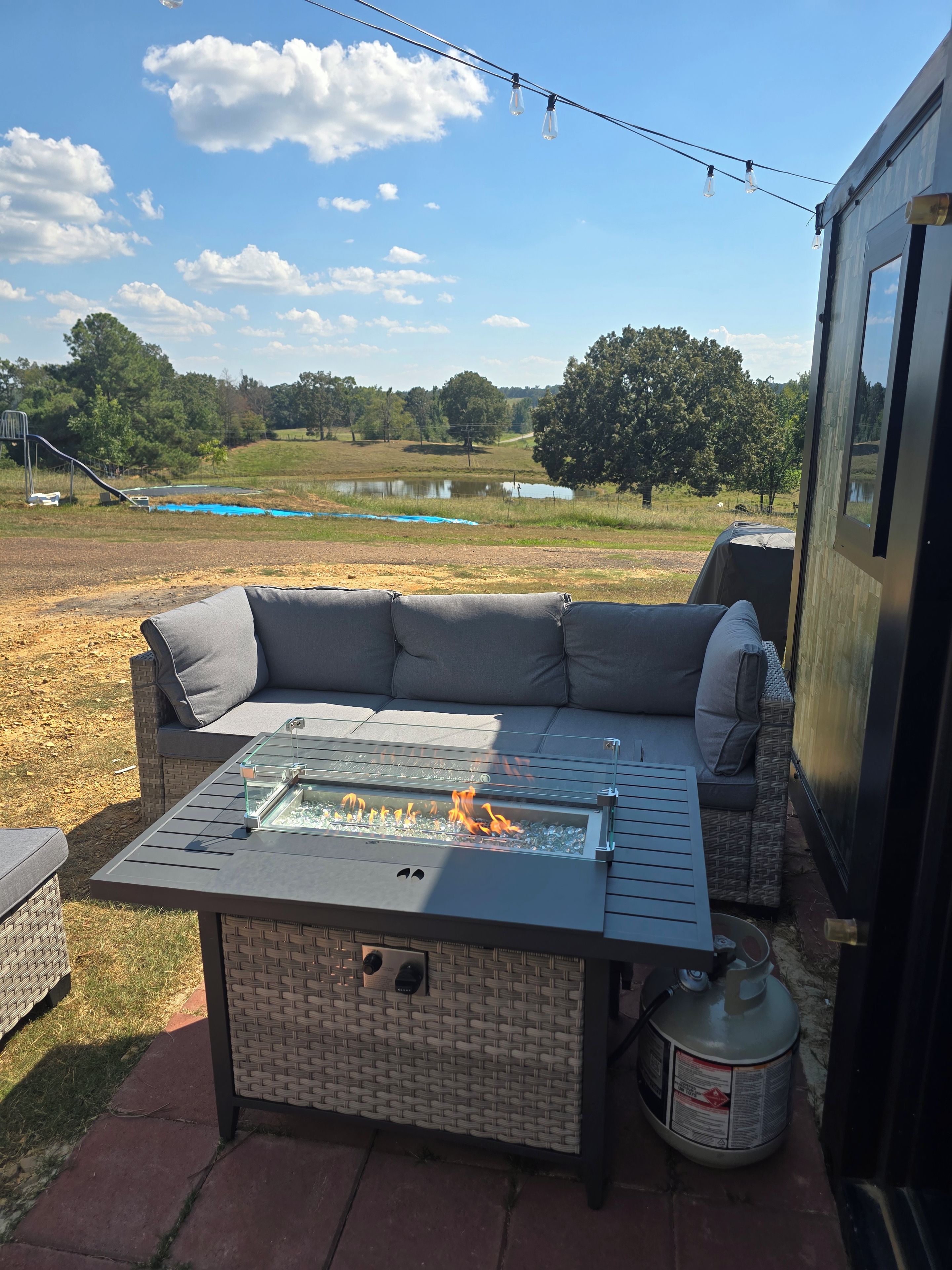Un espace extérieur aménagé avec des sièges, une table à plateau de verre et un foyer, le tout entouré d'un panier en osier, avec vue sur une aire de jeux et des arbres en arrière-plan.
