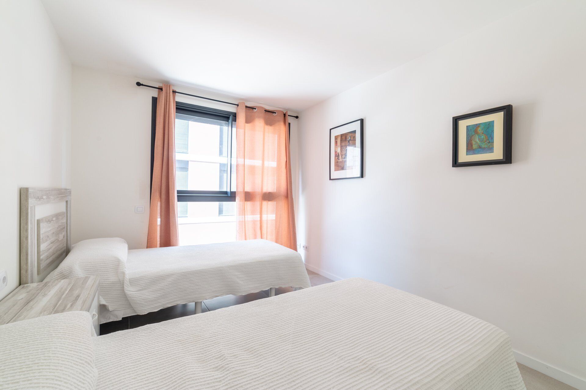 A bedroom with two beds, a window with curtains, and two framed pictures on the wall.