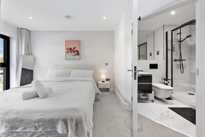 Habitació d'hotel moderna amb un llit gran, una taula de nit, un taulell, un quadre, i un bany amb dutxa, vàter i rentapins.