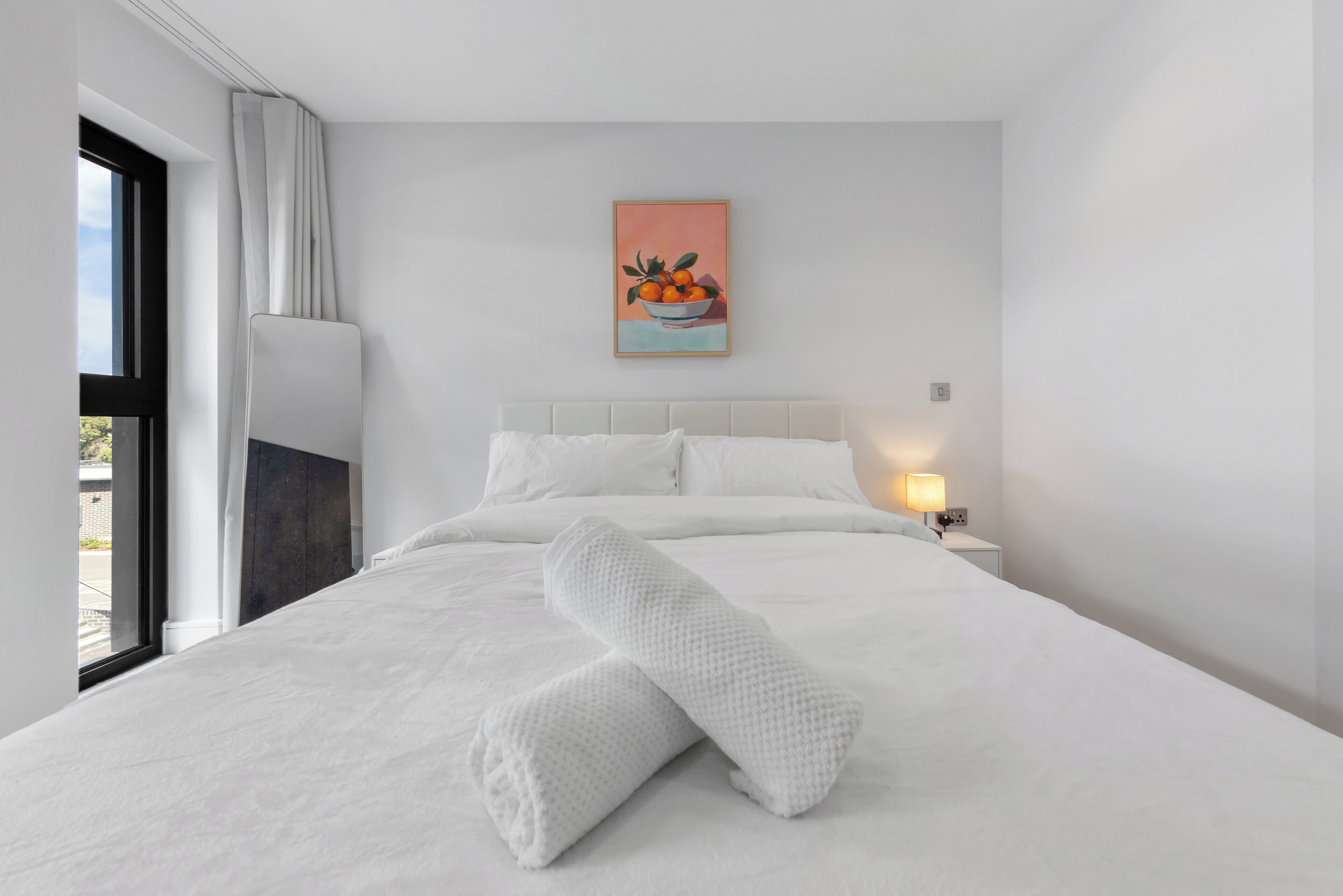 A modern hotel room with a large bed, a nightstand with a lamp, a painting of fruit on the wall, and a window with curtains.
