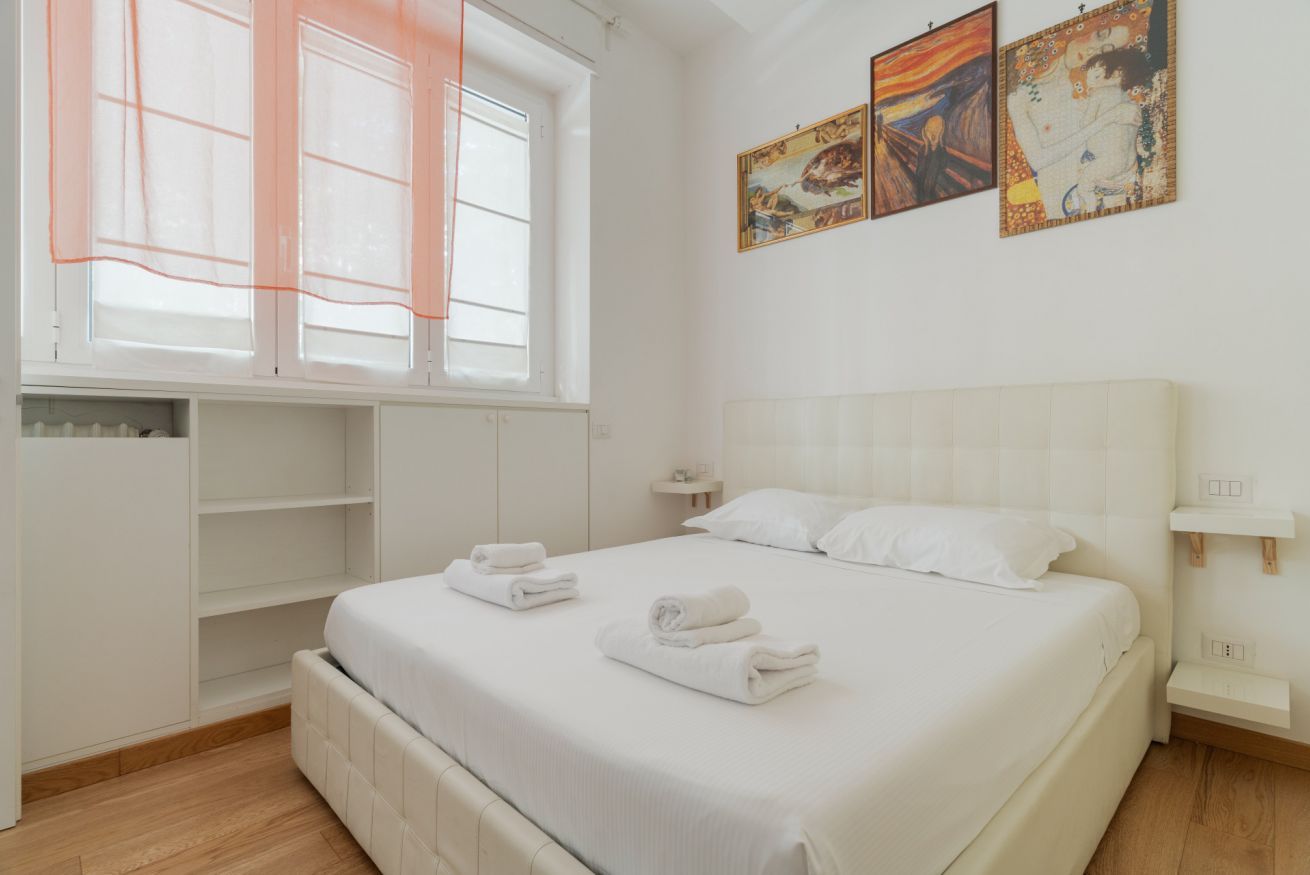 Een slaapkamer met een bed, een raam met zonwering, een nachtkastje en kunst aan de muur.