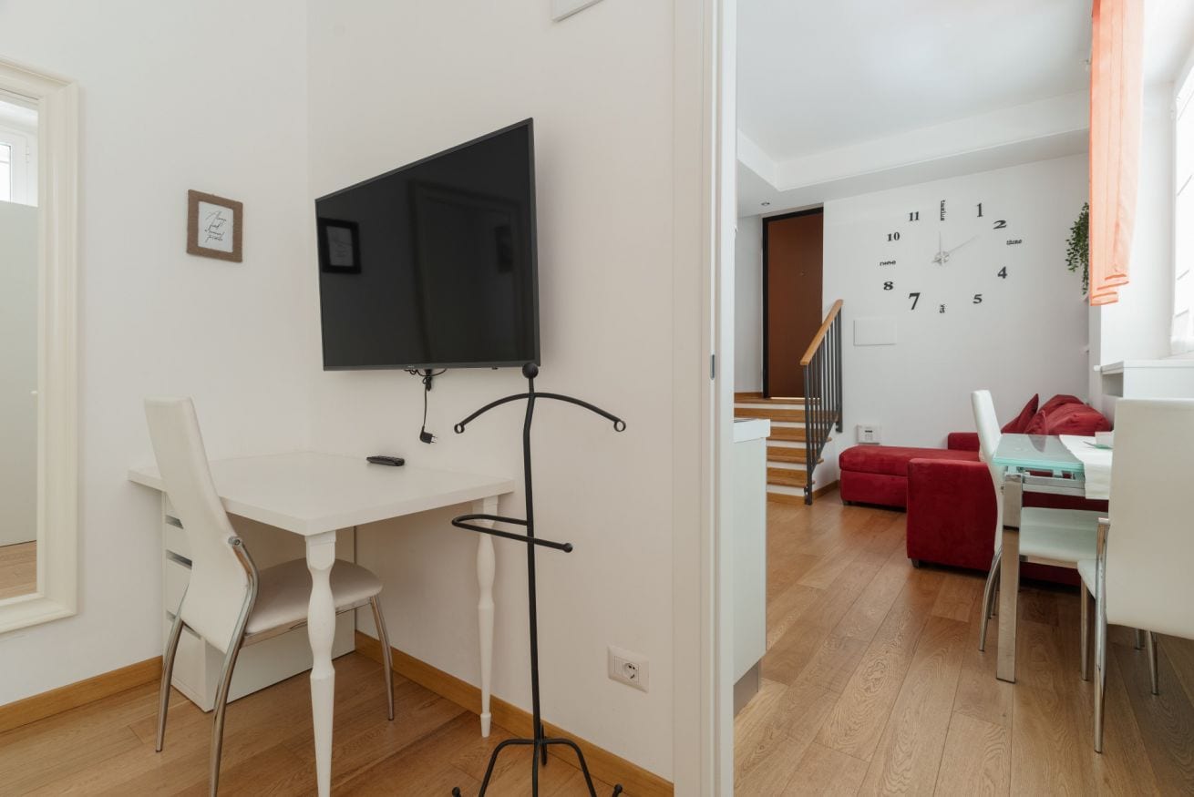 Een moderne woonkamer met een witte bank, een eettafel met stoelen, een aan de muur bevestigde televisie en een trap naar een hoger niveau.