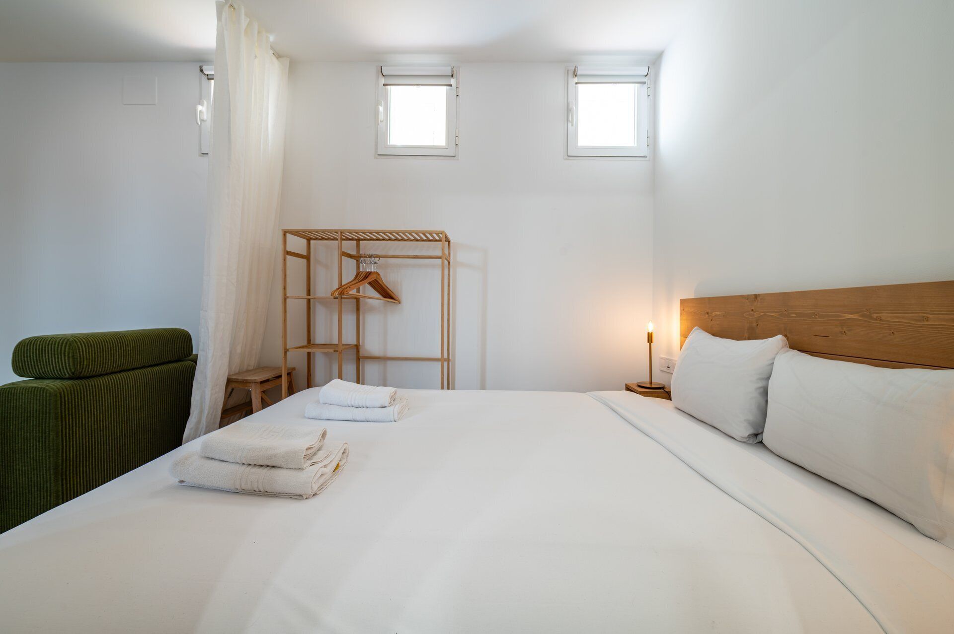 A hotel room with a bed, a wooden headboard, a bedside table with a lamp, a hanging coat rack, and a green chair.