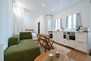 Ein kompakter Wohnraum mit Bett, einem grünen Schlafsofa, einem Holzessttisch, einer Küchenzeile mit Waschmaschine und kleinem Backofen.