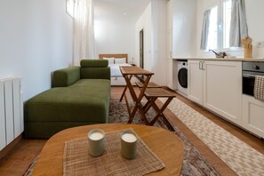 Ein gemütlicher Wohnraum mit einem grünen Sofa, einem Holztisch mit Kerzen, einem Bett und einer Küchenzeile mit Waschmaschine und Backofen.