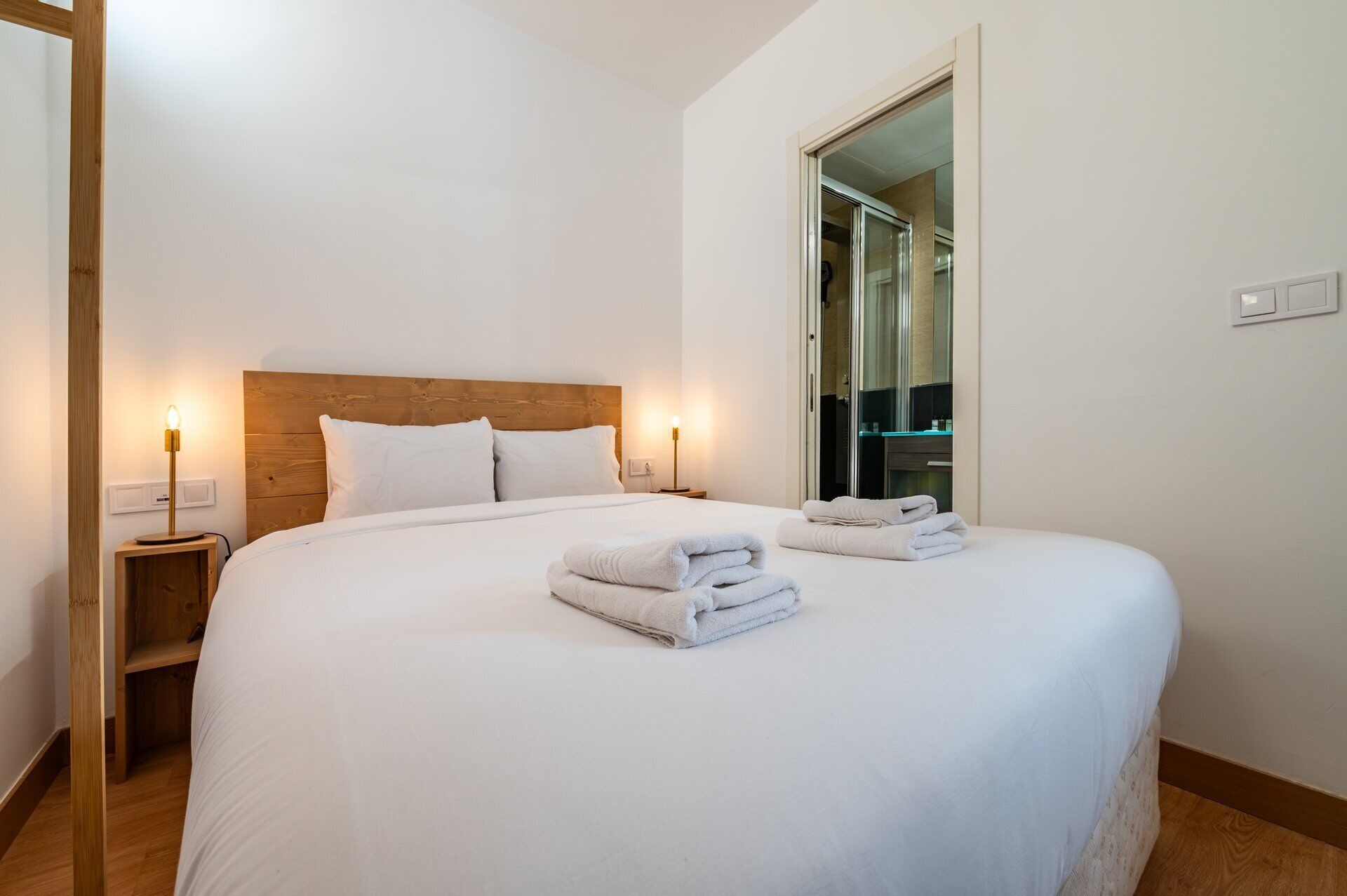 A hotel room with a large bed, two bedside tables with lamps, a wooden headboard, and a bathroom visible through a glass door.