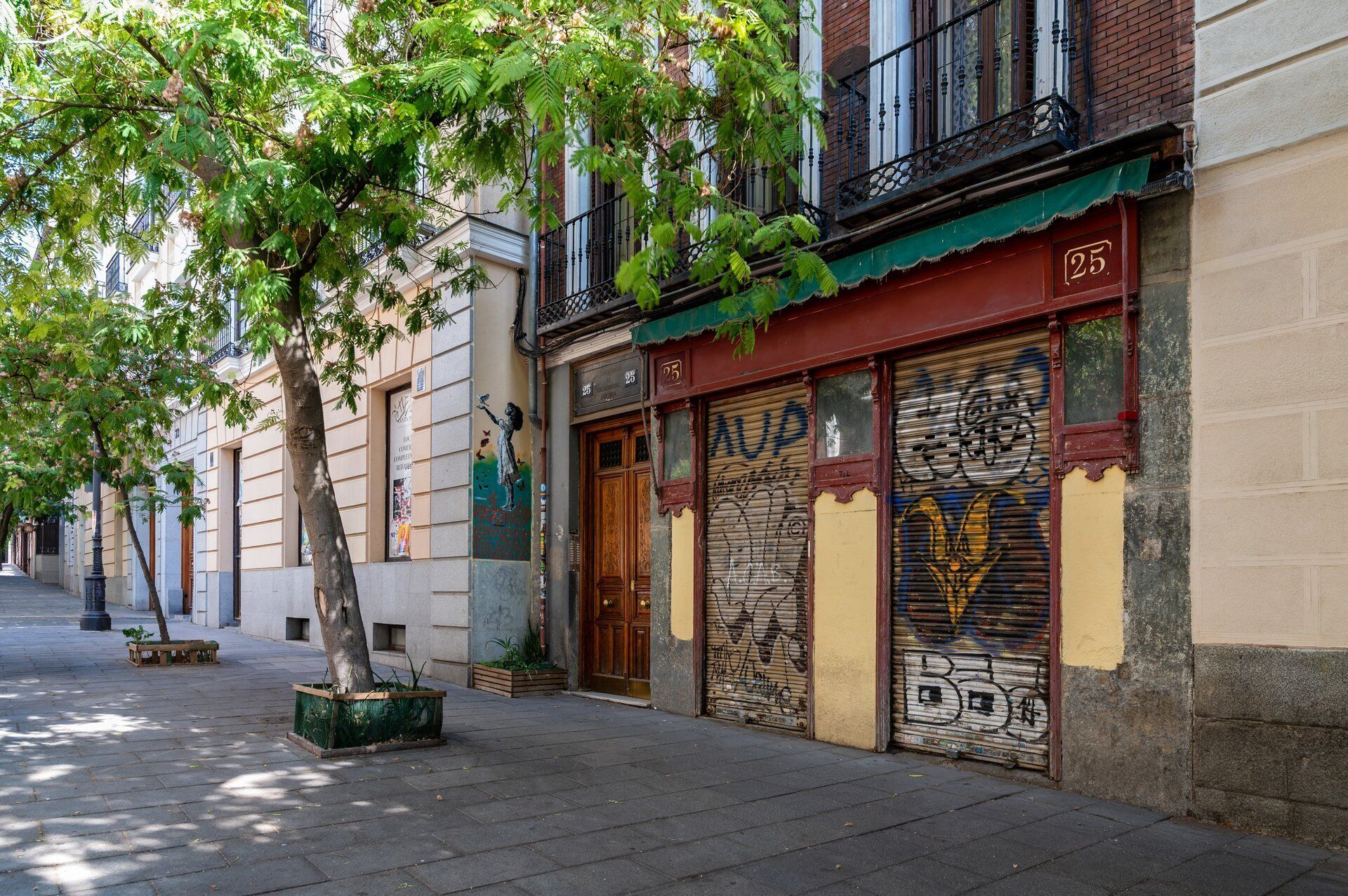 A street with closed shop fronts, a tree-lined sidewalk, and a building with a balcony.