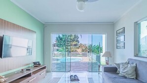 A modern living room with a flat-screen TV, a sofa, and a glass door leading to a pool area. - Casa em Ubatuba com Piscina e Vista para o Mar (Ubatuba)