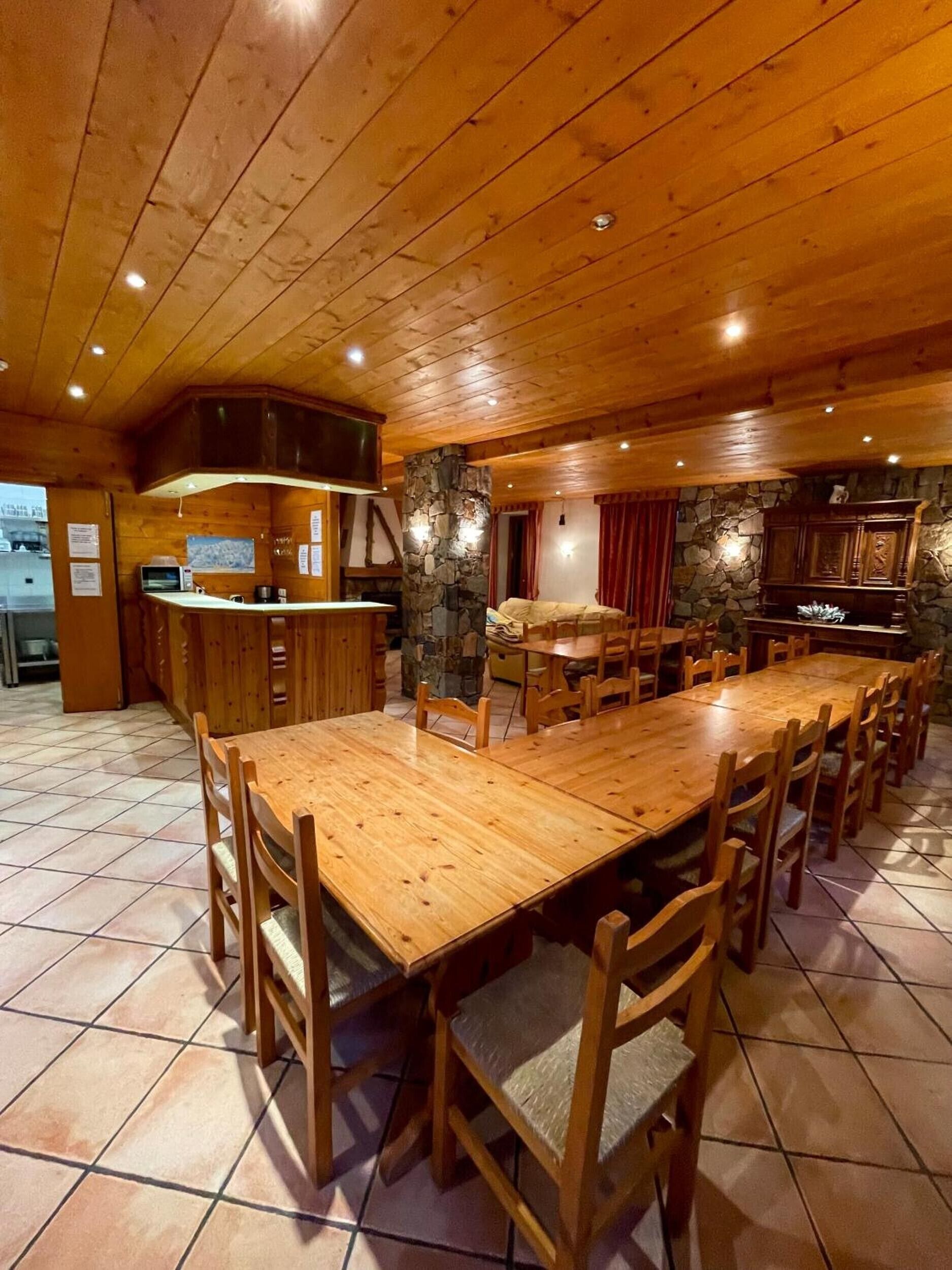 A wooden dining area with a long table and chairs, a kitchen with a stone wall, and a stone fireplace.