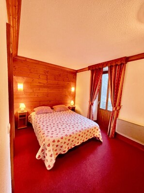 A bedroom with a wooden ceiling, a bed with a patterned bedspread, two bedside lamps, and a window with curtains. - Chalet Le Joly Brides les Bains – 11 Bedrooms, Near Cable Car, Sleeps 23 (Brides-les-Bains)