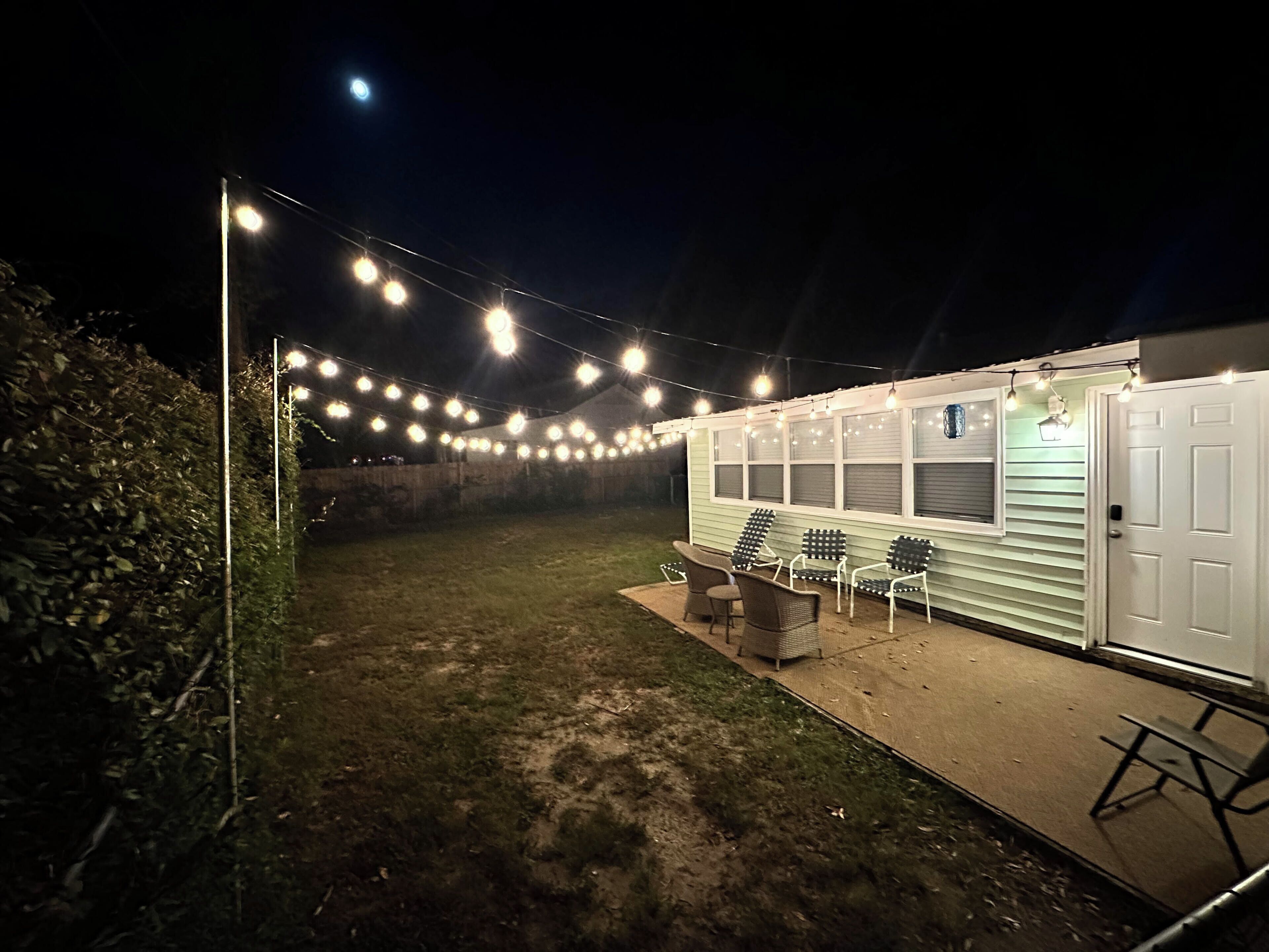 Un espace extérieur abrité, agrémenté de guirlandes lumineuses, un petit coin salon avec des chaises et un bâtiment doté d’une porte.