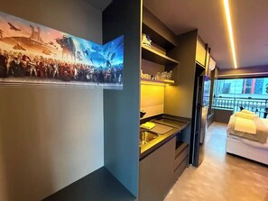 A modern hotel room with a large window, a bed with a yellow towel, a kitchenette, and a wall-mounted TV. - Themed Studio | Liberdade Metro Station (São Paulo)