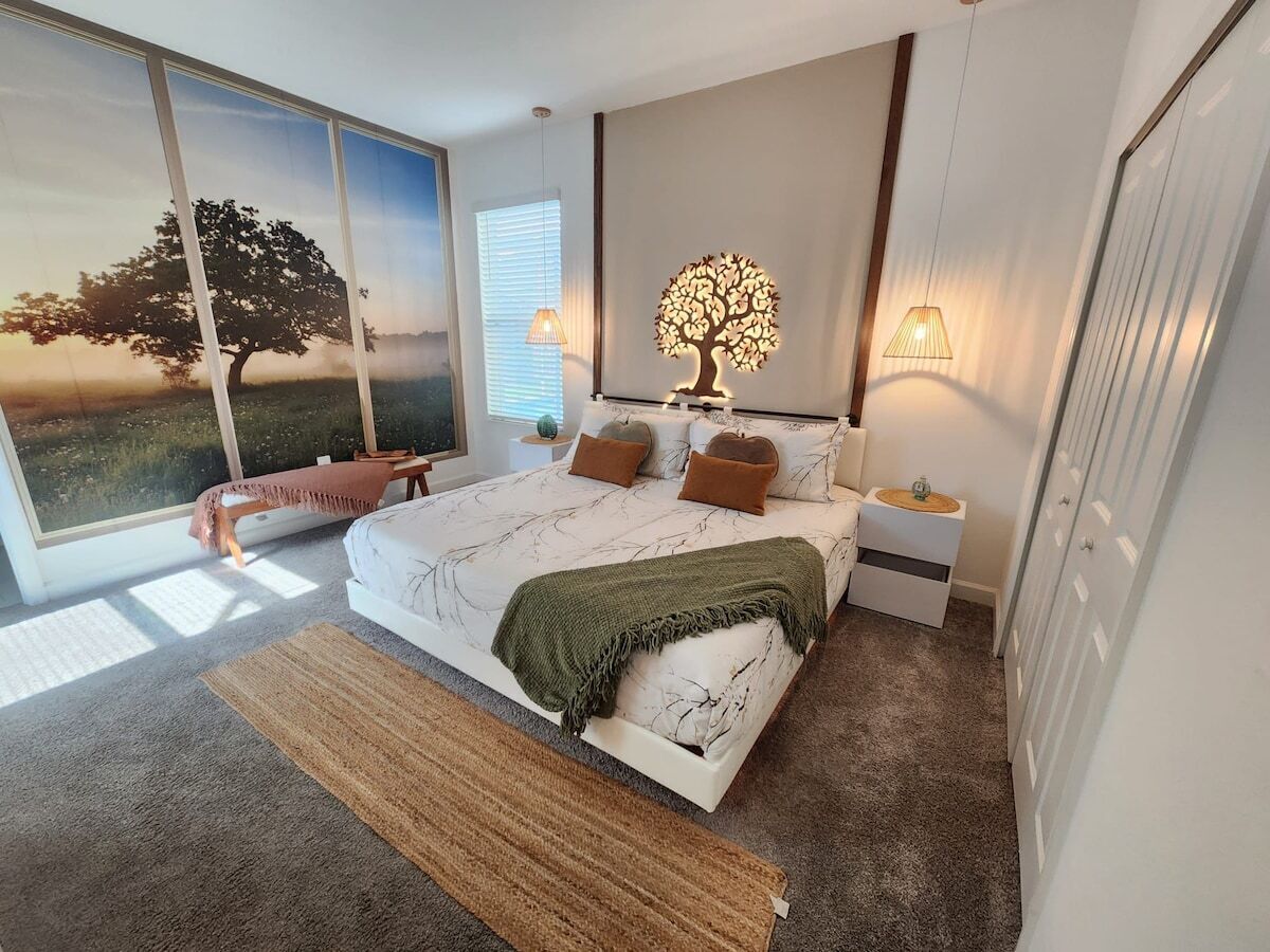 Un dormitorio moderno con una cama grande, una mesita de noche, una lámpara de pared con forma de árbol y vistas a un árbol a través de una ventana.