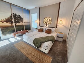 A modern bedroom with a large bed, a nightstand, a wall-mounted tree light, and a view of a tree through a window. - Villa w/ Private Pool, Near Disney - Champions (Davenport)