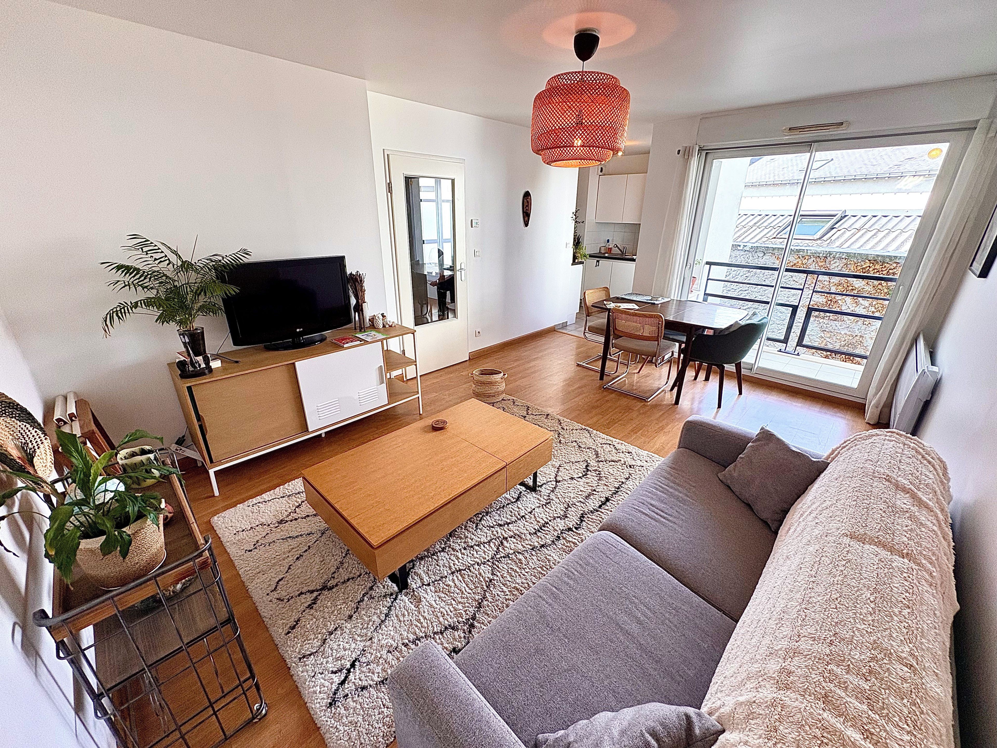 A modern living room with a sofa, coffee table, and TV. There's a dining area with chairs and a table. A large window provides natural light and a view of the outside.