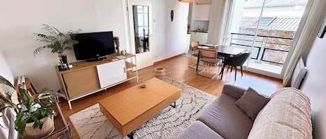 Un salon moderne avec un canapé, une table basse et un téléviseur. Il y a également un coin repas avec des chaises et une table. Une grande fenêtre laisse entrer la lumière naturelle et offre une vue sur l’extérieur.