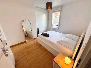 Ein Schlafzimmer mit Bett, Nachttisch mit Lampe, Spiegel und Fenster mit Blick auf Gebäude.