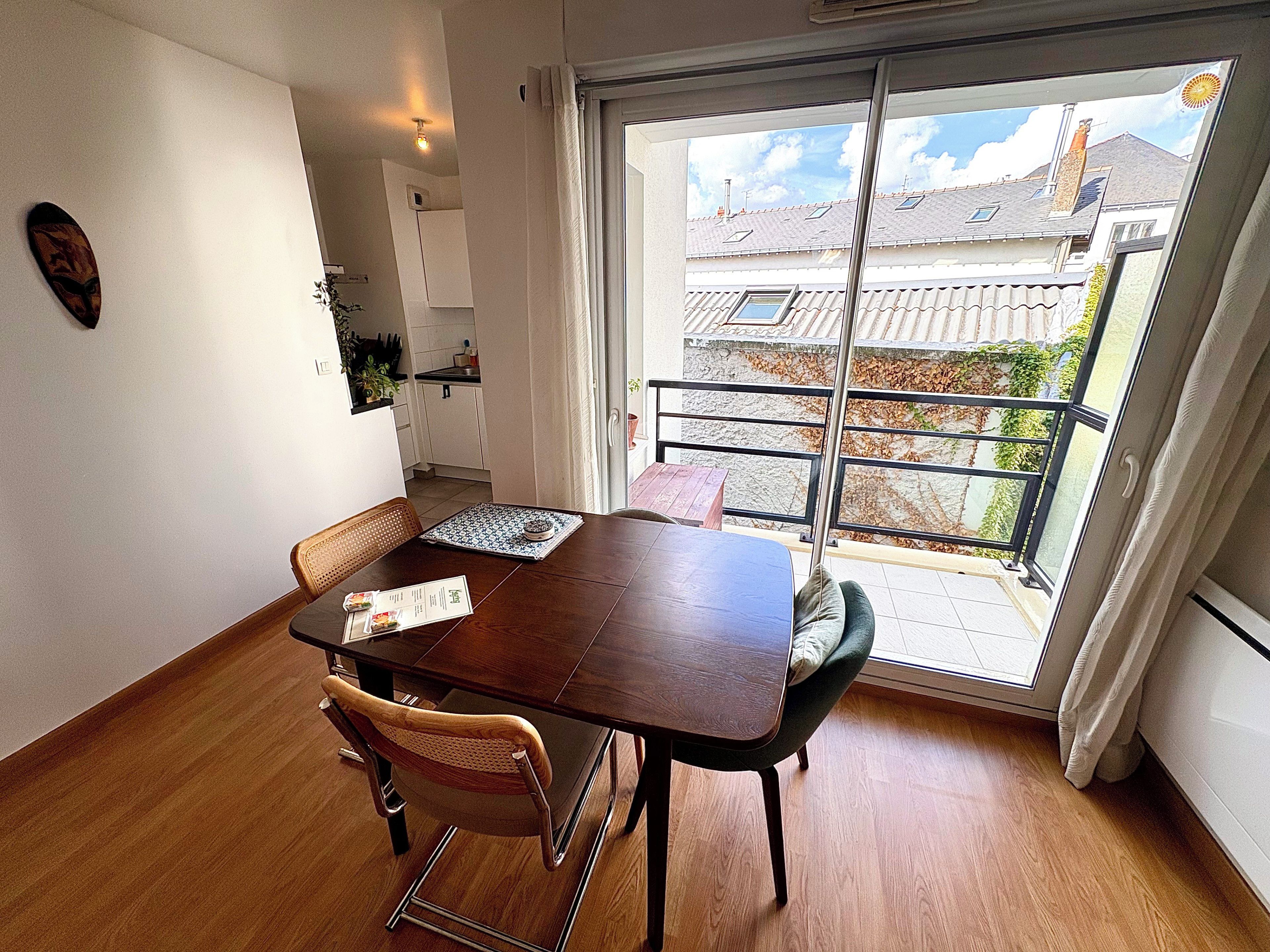Un coin repas avec une table et des chaises en bois, une vue sur l’extérieur grâce à une porte coulissante, et une cuisine en arrière-plan.